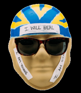 Unmasking Brain Injury mask painted with yellow and blue on the forehead, large sunglasses and text
