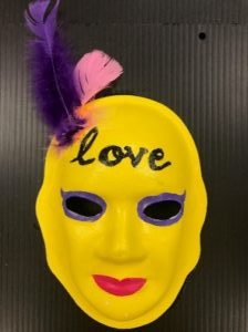 Unmasking Brain Injury mask painted yellow with a purple and pink feather and "love" painted on the forehead