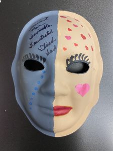 Unmasking Brain Injury mask painted half yellow and half blue with hearts and text