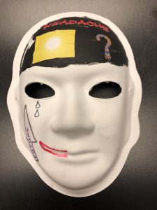 Unmasking Brain Injury mask painted with black on the forehead and a yellow square and "headache" on the forehead