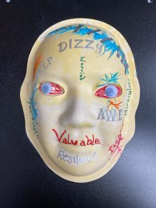 Unmasking Brain Injury mask painted yellow with words and painted bloodshot eyes