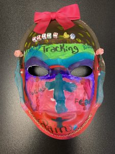 Unmasking Brain Injury mask painted with red, blue, purple, green and black sections and text, a bow and beads