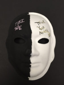 Unmasking Brain Injury mask painted half black and half white with text