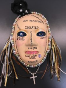 Unmasking Brain Injury mask painted with text and with hair and a cross necklace