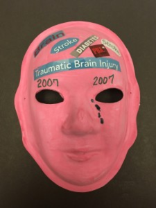 Unmasking Brain Injury mask painted red with the text "2007" and magazine cut outs collaged on it