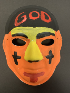Unmasking Brain Injury mask painted orange, black and yellow with text "God" on forehead and crosses on cheeks