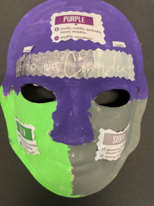 Unmasking Brain Injury mask painted purple, green and gray with text explaining the color choices