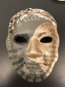 Unmasking Brain Injury mask painted with gold and sage green with text and ornamentation