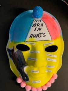 Unmasking Brain Injury mask painted in blue, red, yellow and black and with the words "my brain hurts"