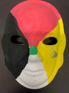 Unmasking Brain Injury mask painted with black, red, yellow and white sections and a green spot and sparkly tear