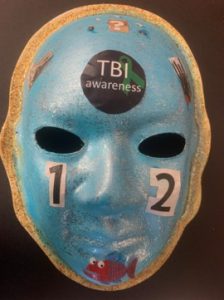 Unmasking Brain Injury mask painted with sparkly blue paint, a fish and a TBI awareness sticker